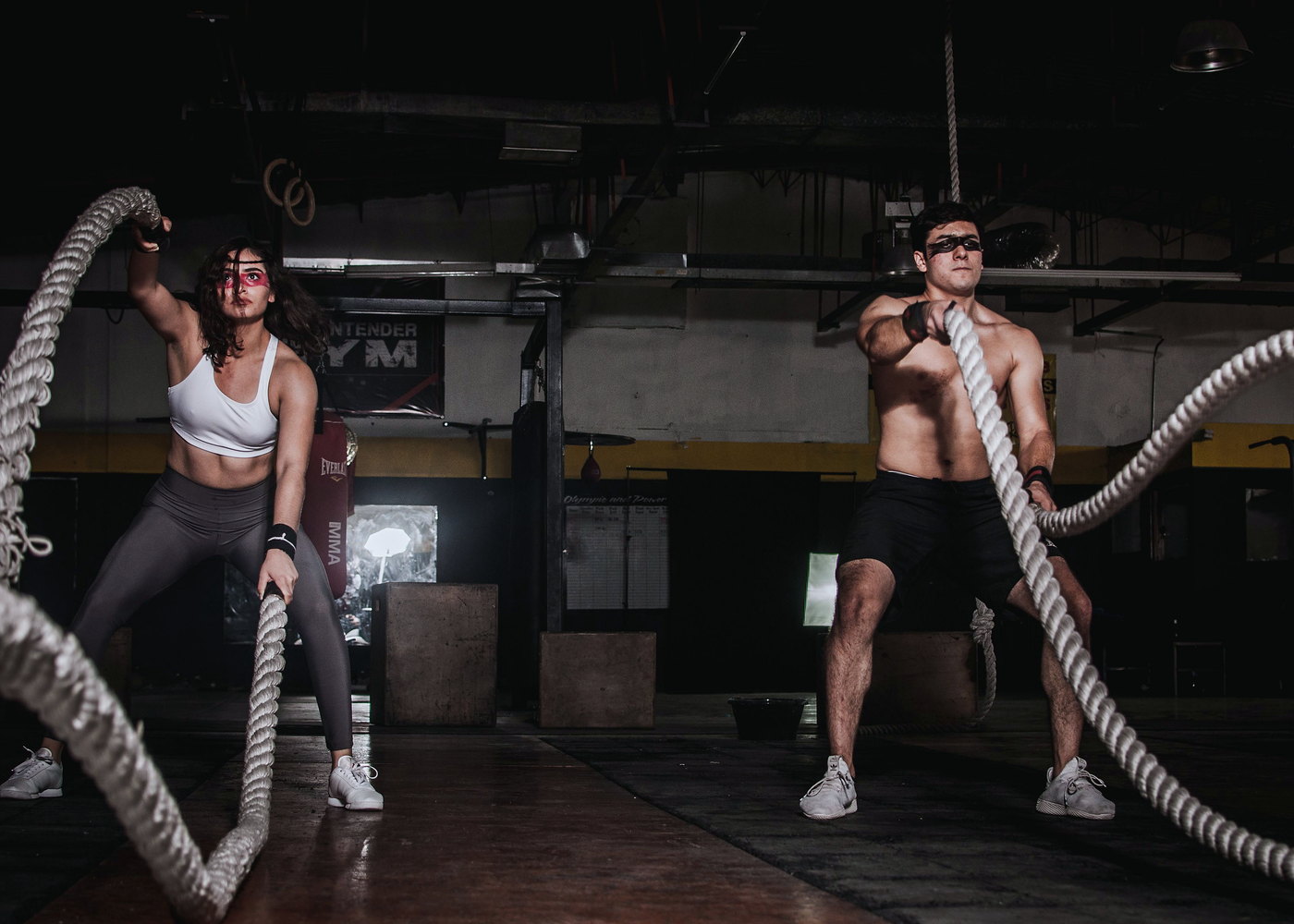 Two people using battle ropes in a gym with hanging rings and boxing equipment.